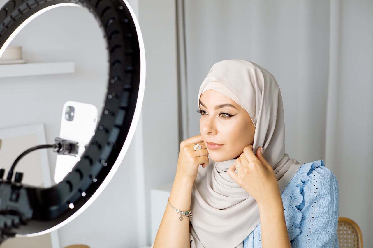 Woman in hijab using smartphone and ring light for video recording indoors.